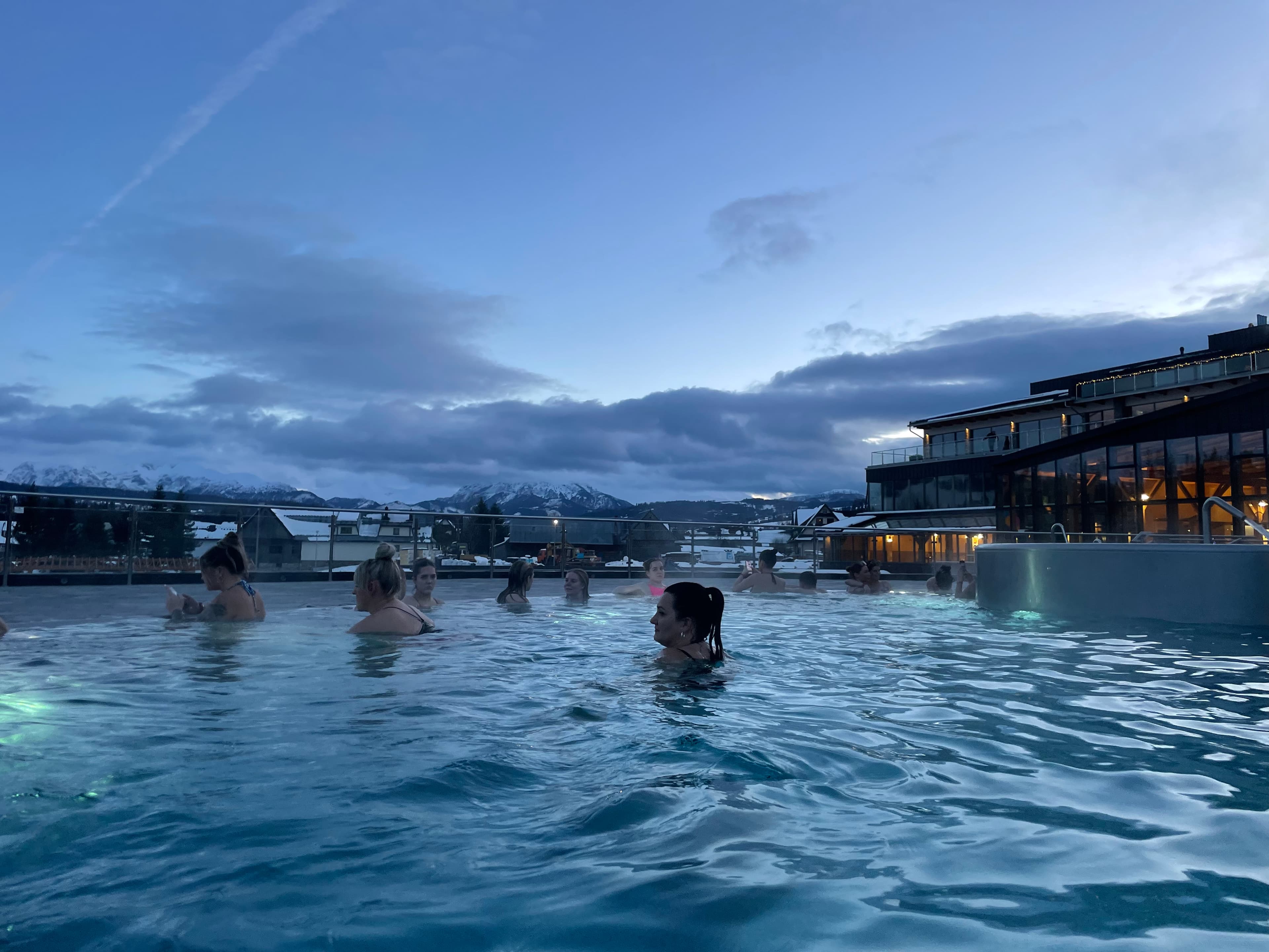 People relaxing in an outdoor pool at dusk with mountains and buildings in background.